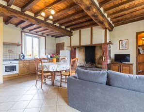 A cozy living room with a fireplace, wooden beams, a dining area with chairs, and a kitchen with cabinets and a window. - House with Garden, Pets allowed (Hauteville-la-Guichard)