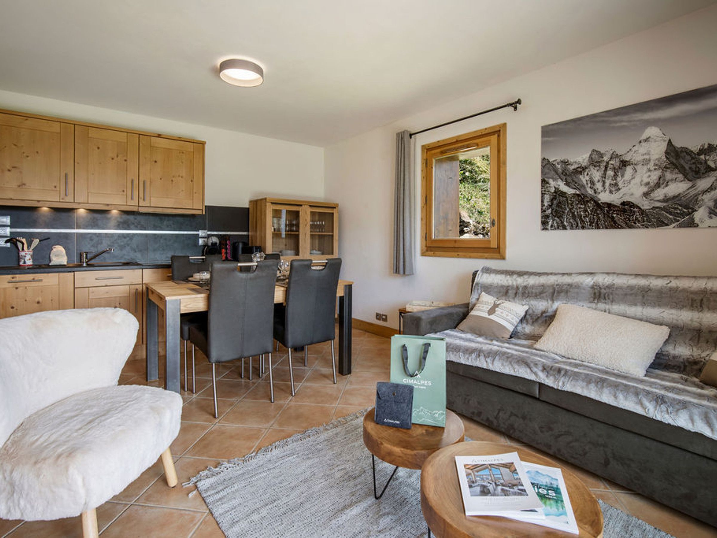 Ein modernes Wohnzimmer mit einer Couch, einem Essbereich mit Stühlen, einer Küche mit Holzschränken und einem großen Fenster mit Blick auf die Berge.