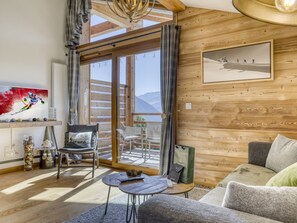 A cozy room with a wooden interior, a sofa, a chair, a table, and a large window offering a view of the mountains. - 2-bedroom cabin apartment for 6 with garage in Les Saisies (Les Saisies)