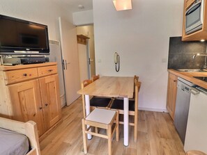 A compact kitchen with wooden cabinets, a dining table, and chairs. A television is mounted above the cabinets.