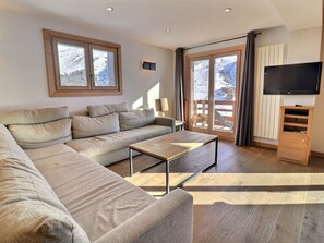 A modern living room with a large beige sectional sofa, a wooden coffee table, a flat-screen TV, and a sliding glass door leading to a balcony with a view of snow-covered mountains.