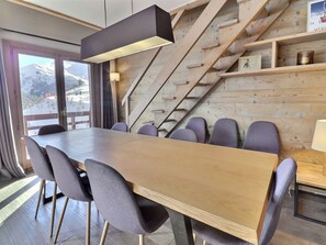 A modern dining area with a long wooden table and grey upholstered chairs, a staircase with wooden steps, and a view of a snowy mountain through large windows.