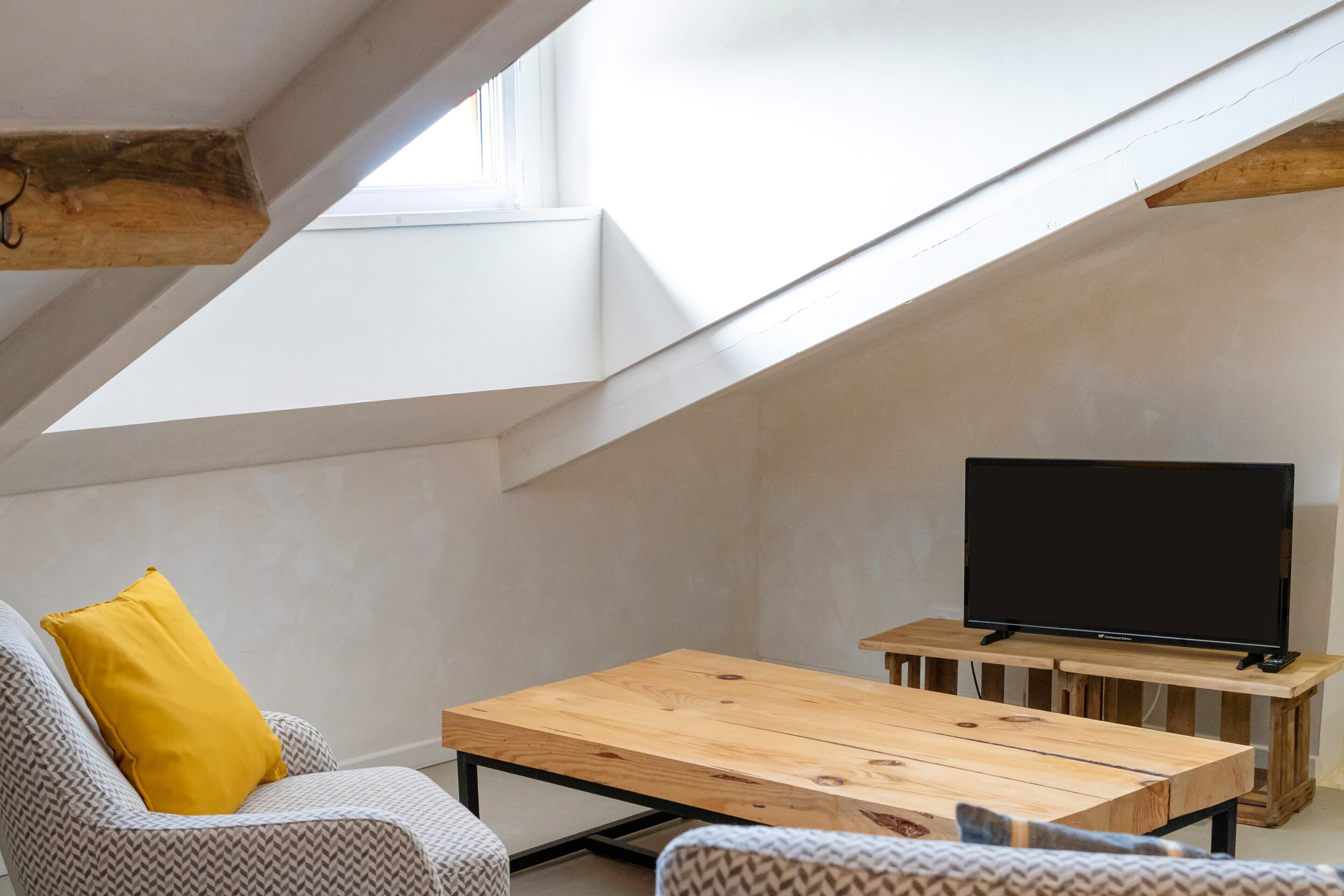 Un dormitorio estilo loft moderno con techo inclinado, una mesa de comedor de madera, un sofá con un cojín amarillo y un televisor sobre un mueble de madera.