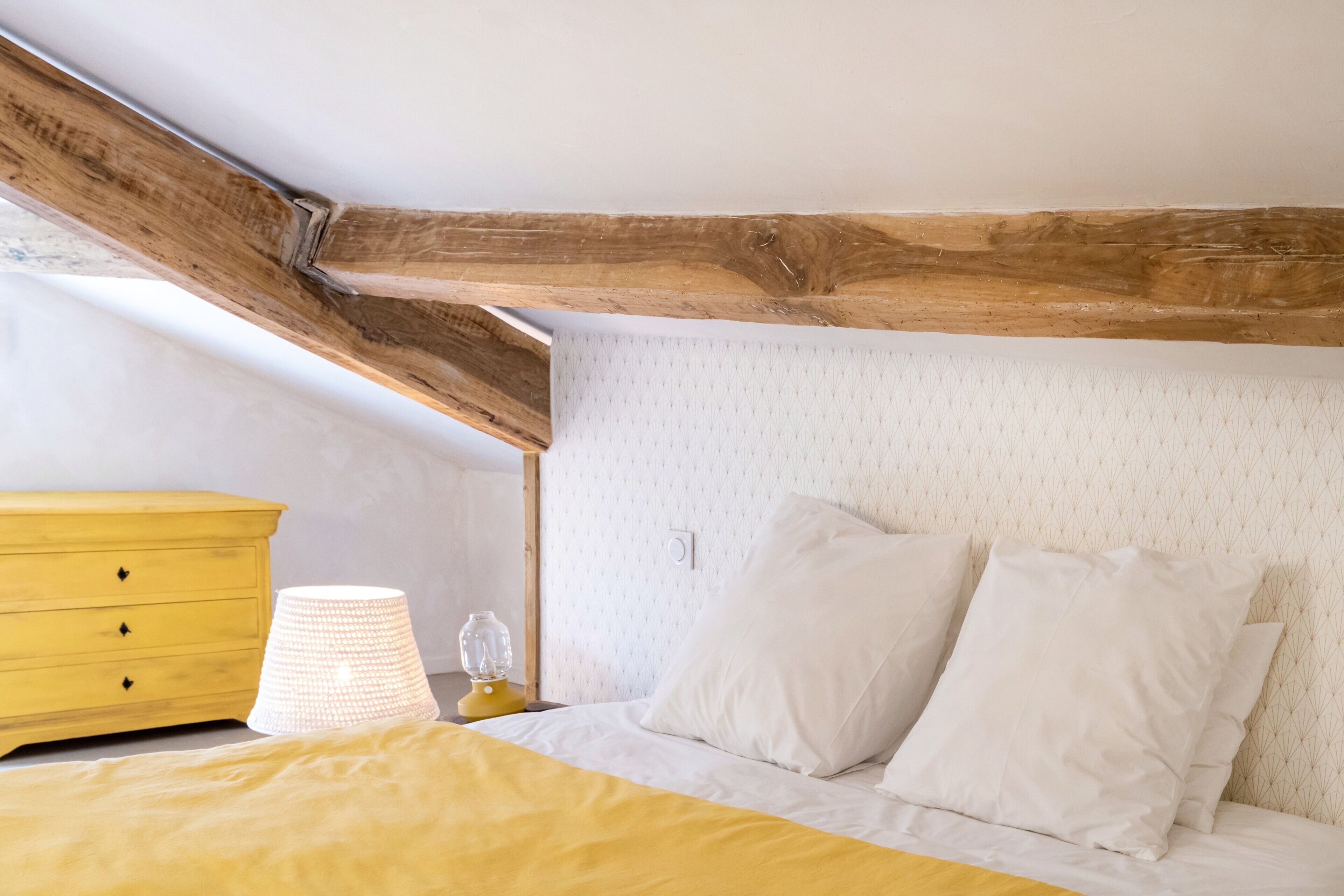 A bedroom with a bed, a nightstand, a lamp, a dresser, and exposed wooden beams.