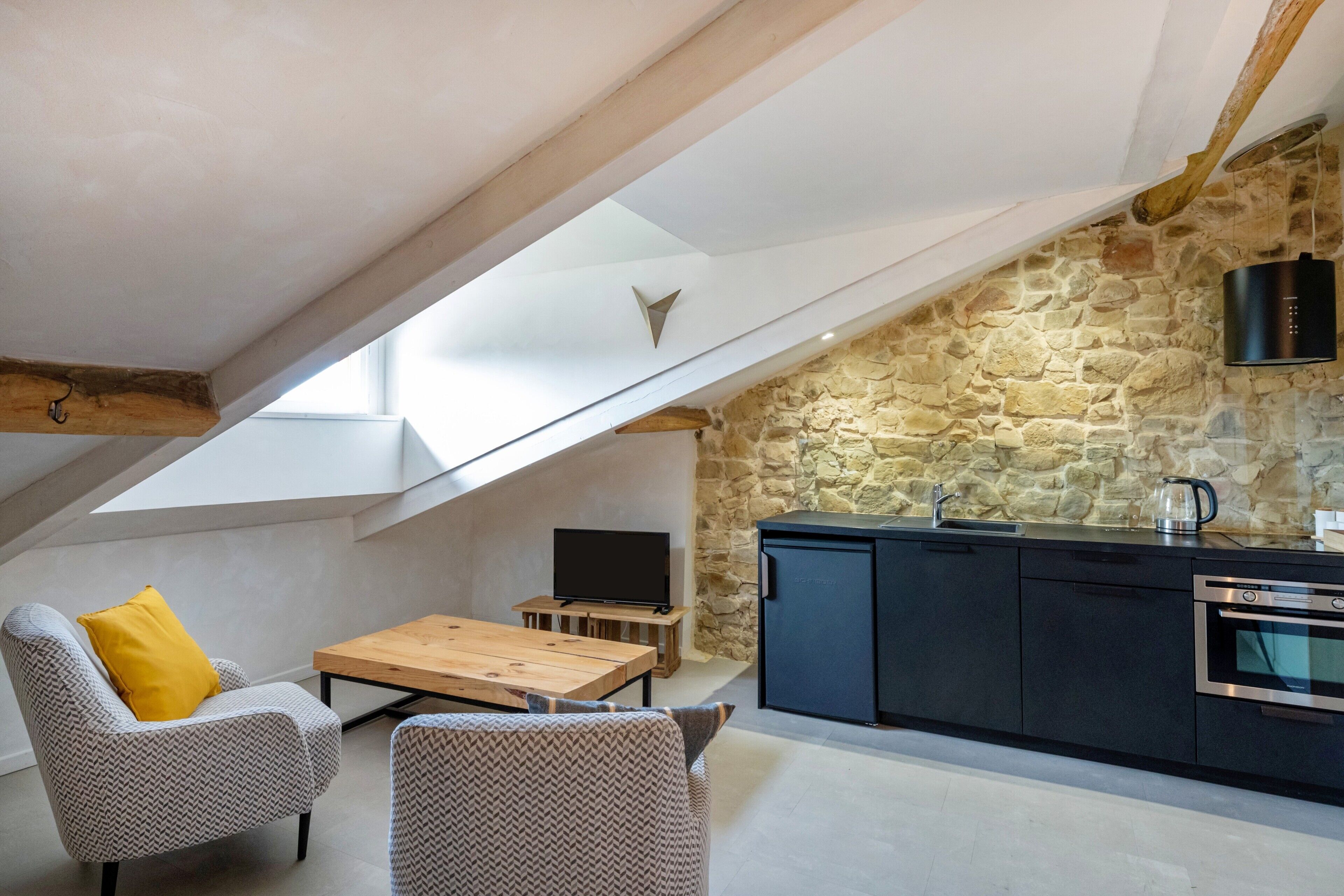 Een loft-achtige kamer met een stenen muur, een houten eettafel, twee gestreept stoelen, een zwart keukenhoekje met een kookplaat en een tv op een standaard.