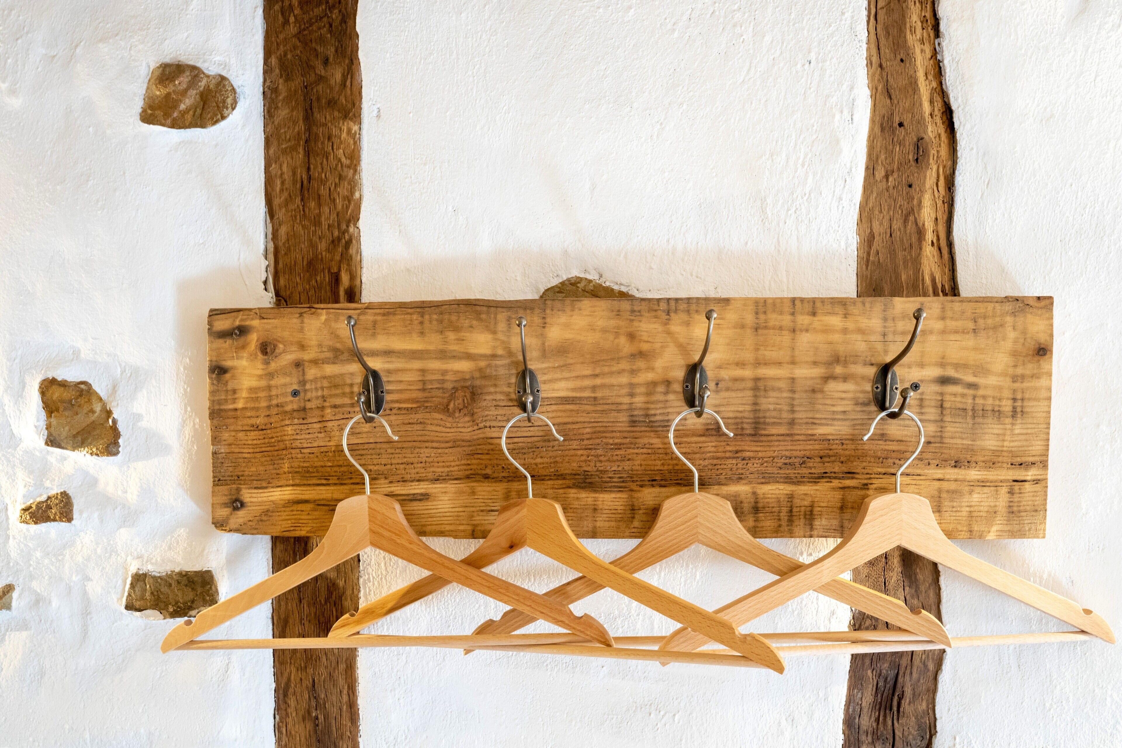 Ein hölzerner Kleiderhaken mit vier Haken und drei Holzaufhängern vor einer weißen Wand mit freiliegenden Holzbalken.