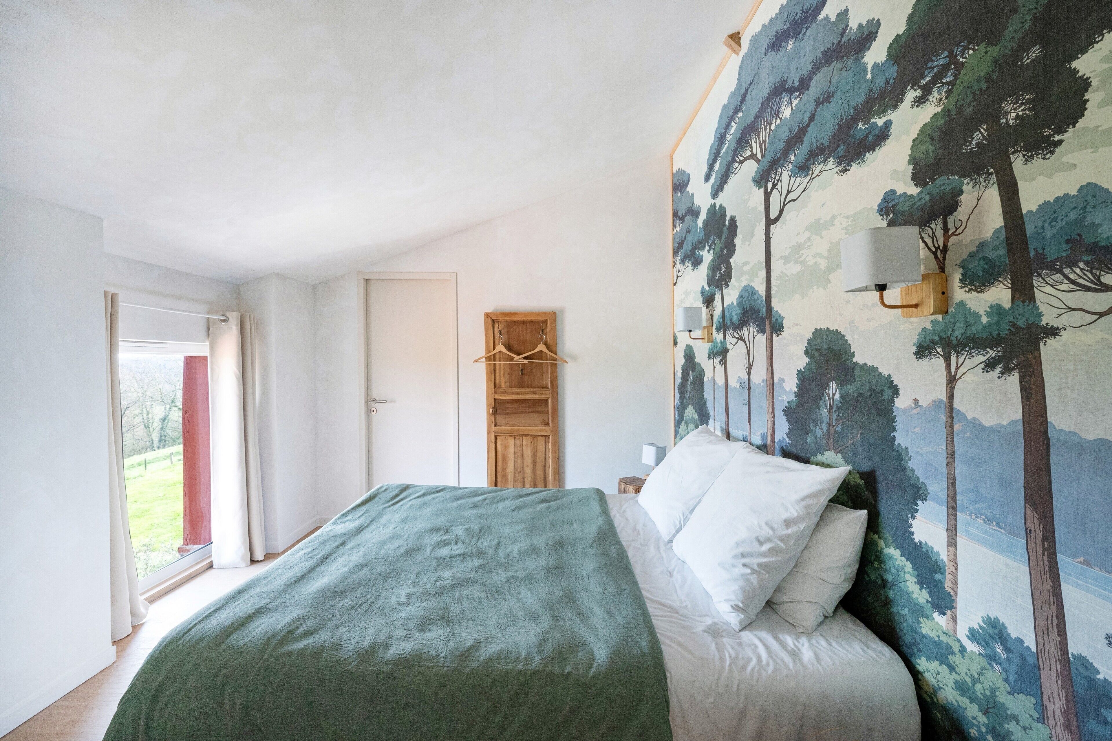 A bedroom with a bed, a wooden wardrobe, a hanging coat rack, and a wall with a forest mural.