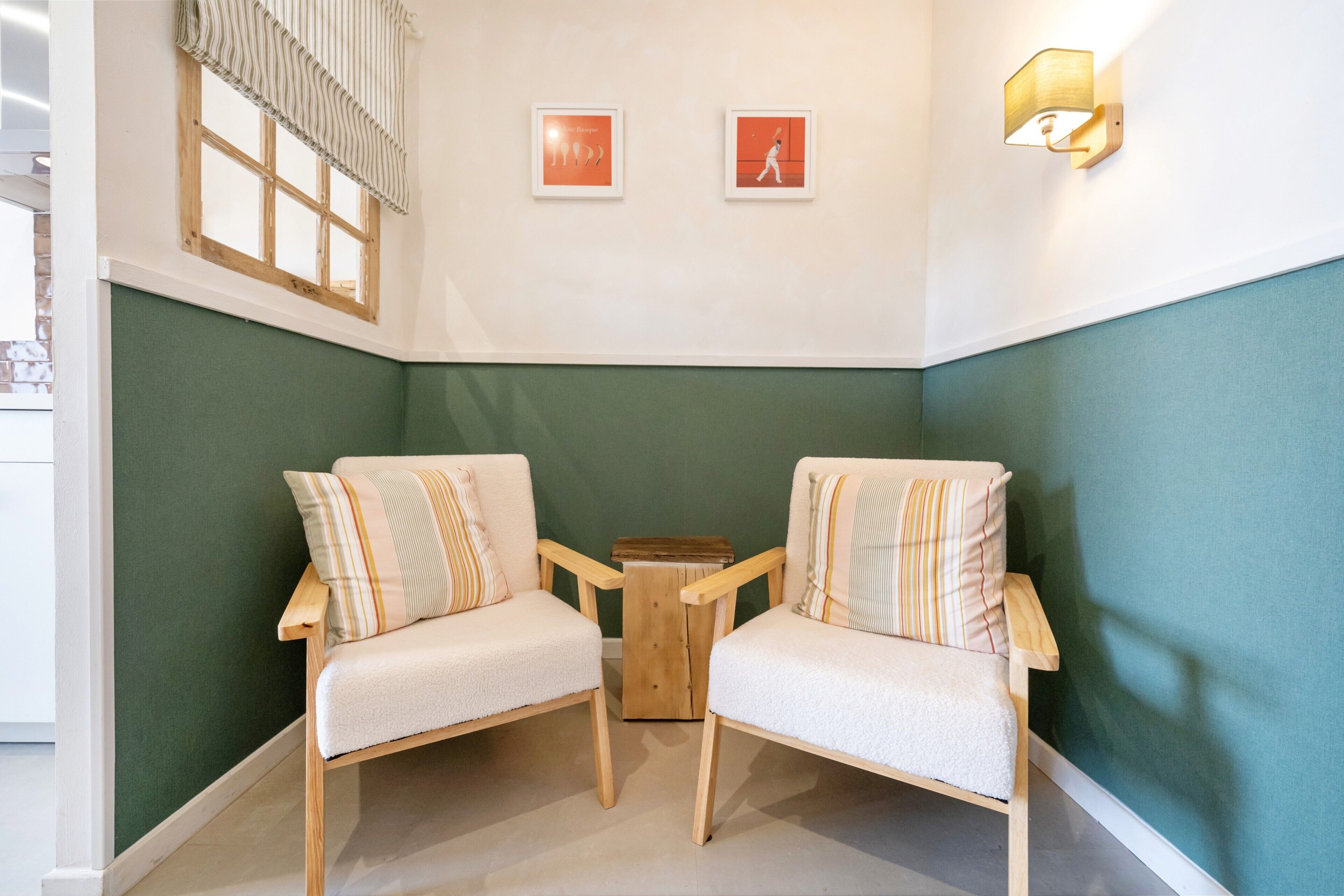 Une pièce d’angle avec deux chaises, une petite table, une fenêtre avec des rideaux et deux tableaux encadrés accrochés au mur.