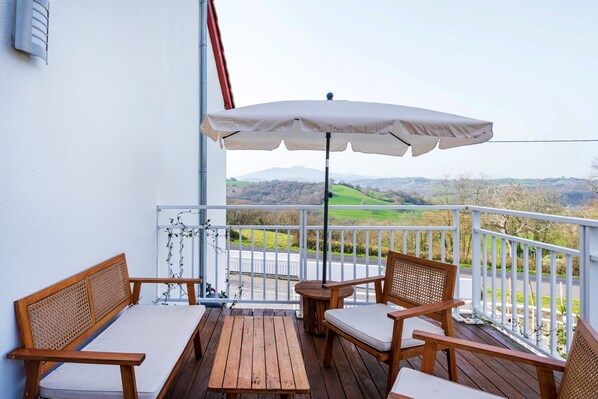 Eine Holzterrasse mit Tisch, zwei Stühlen und Sonnenschirm, mit Blick auf grüne Felder und Hügel.