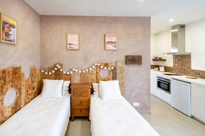 Ein Schlafzimmer mit zwei Betten, einem Holzkopfstück mit Beleuchtung, einer kleinen Kommode und einer Küchenzeile inklusive Herd und Spüle.