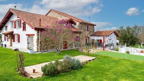 Una casa de piedra de dos pisos con techo de tejas rojas, un jardín con terraza de madera y un pequeño estanque.