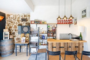 Zona de bar con estantes llenos de botellas, un dardo y una barra de madera con sillas.