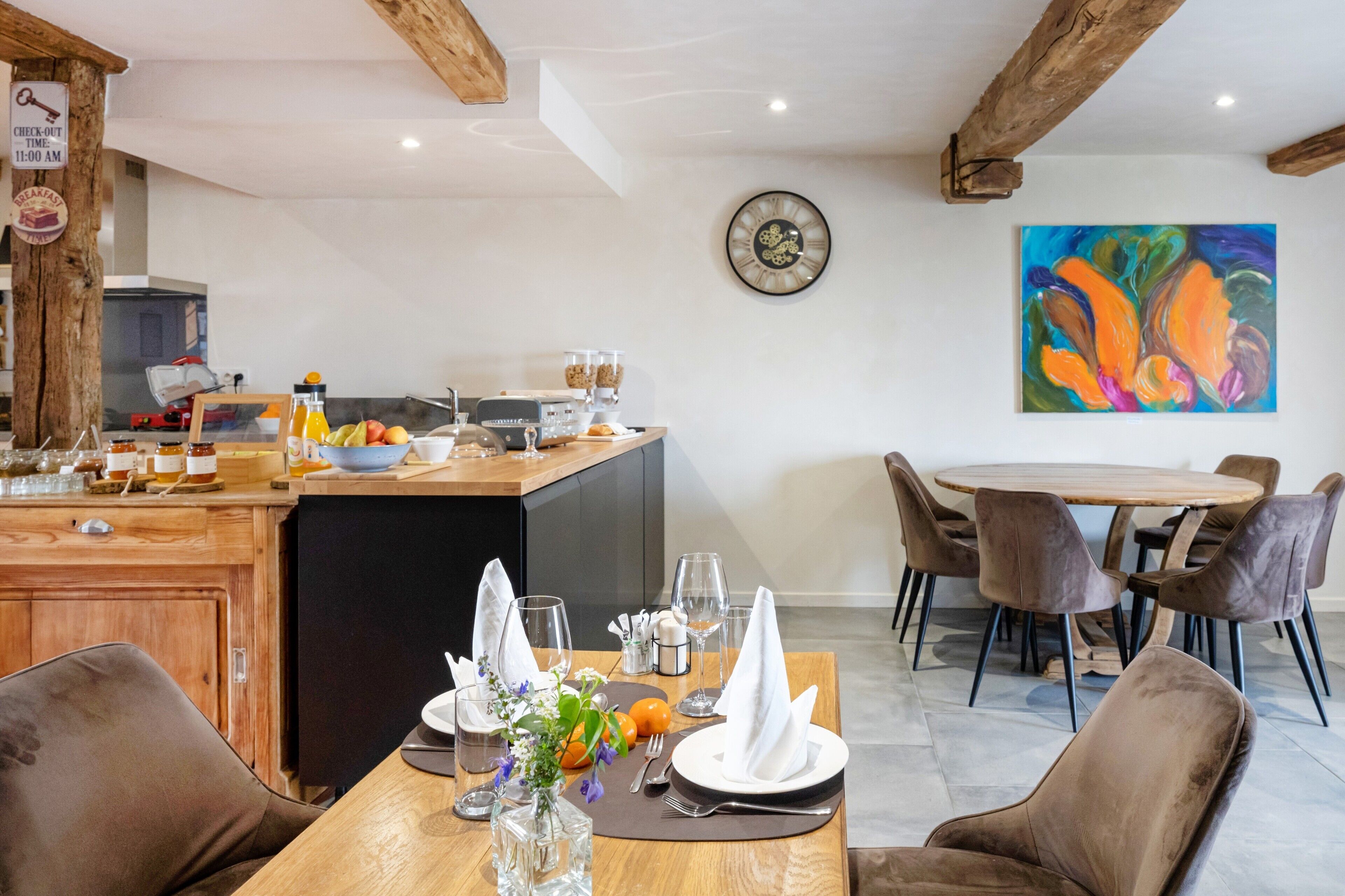 Uma sala de jantar com uma mesa de madeira posta para dois, cozinha ao fundo e um quadro abstrato colorido na parede.