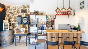 Zona de bar con estantes llenos de botellas, un dardo y una barra de madera con sillas.