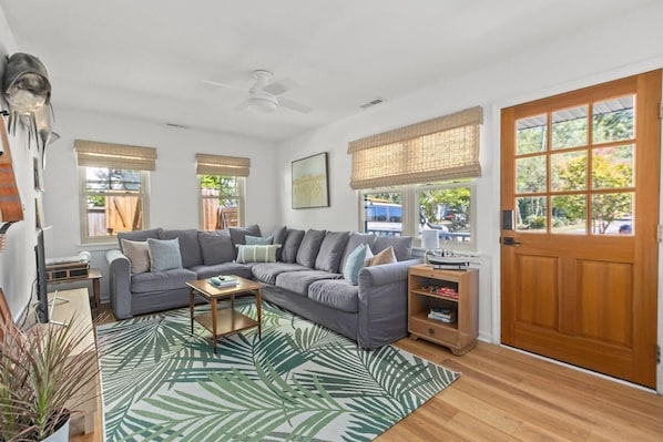 A living room with a grey sectional sofa, a coffee table, a wooden side table, and a large window with a view of the outdoors. - Shem Shack-Old Village, 5 mins to Beach (Mount Pleasant)