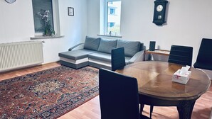A modern living room with a sectional sofa, a dining table with chairs, a large rug, and two clocks on the walls. - 2-room apartment in the center of Friedrichsdorf (Friedrichsdorf)
