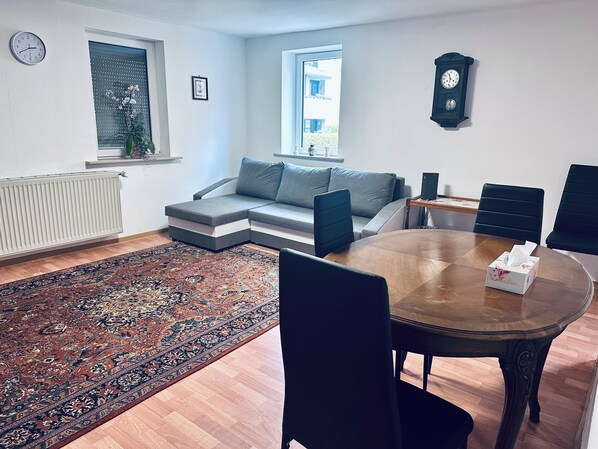 A modern living room with a sectional sofa, a dining table with chairs, a large rug, and two clocks on the walls. - 2-room apartment in the center of Friedrichsdorf (Friedrichsdorf)