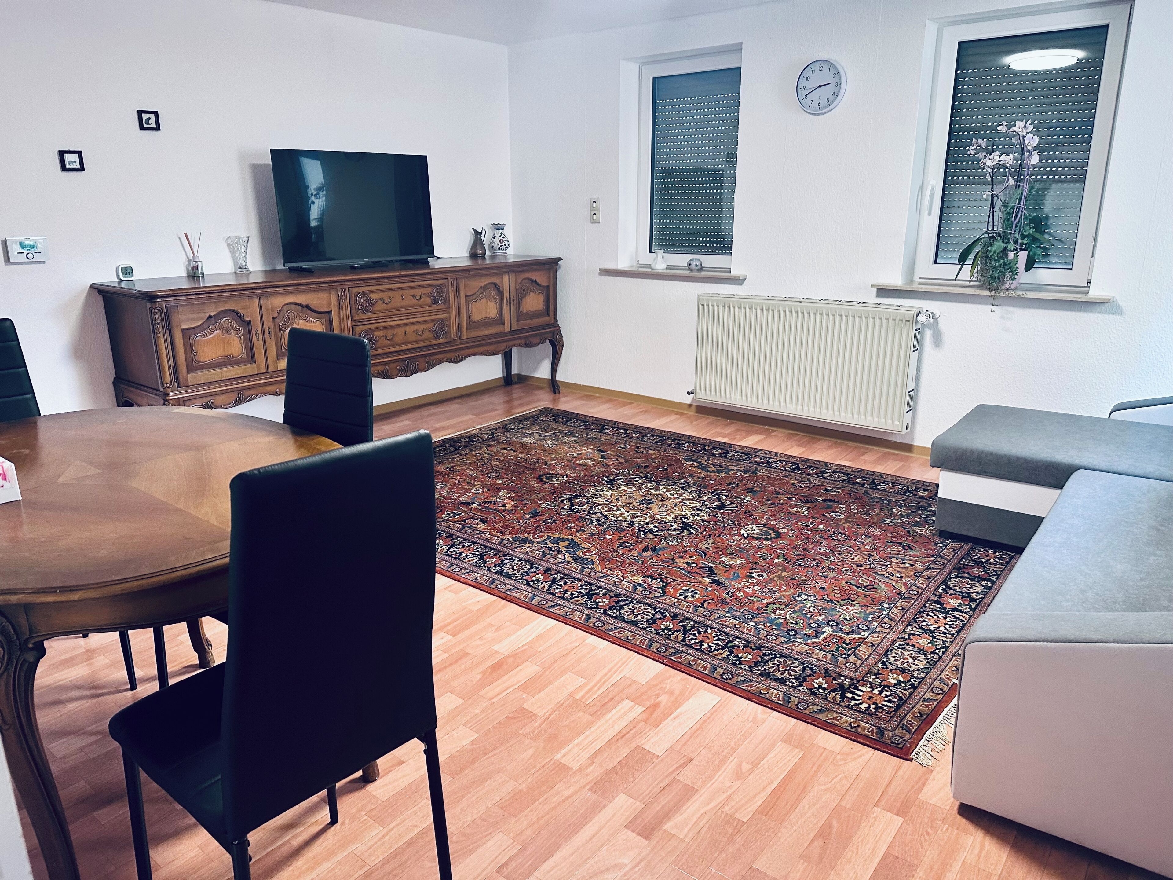 Een woonkamer met een houten eettafel, stoelen, een televisie op een houten kast, een groot kamerkleed, een grijze bank en een raam met zonwering.