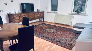 Een woonkamer met een houten eettafel, stoelen, een televisie op een houten kast, een groot kamerkleed, een grijze bank en een raam met zonwering.