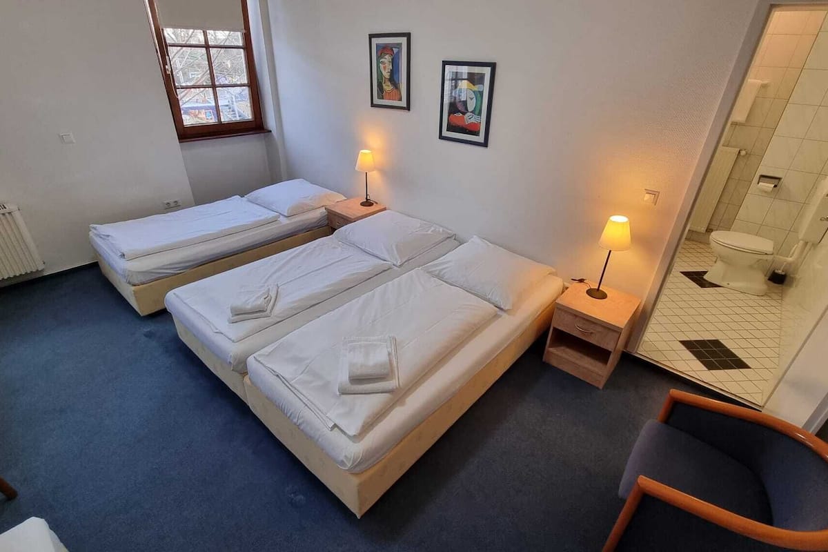 A hotel room with two single beds, a small desk with a lamp, a chair, and a bathroom visible through a glass door.