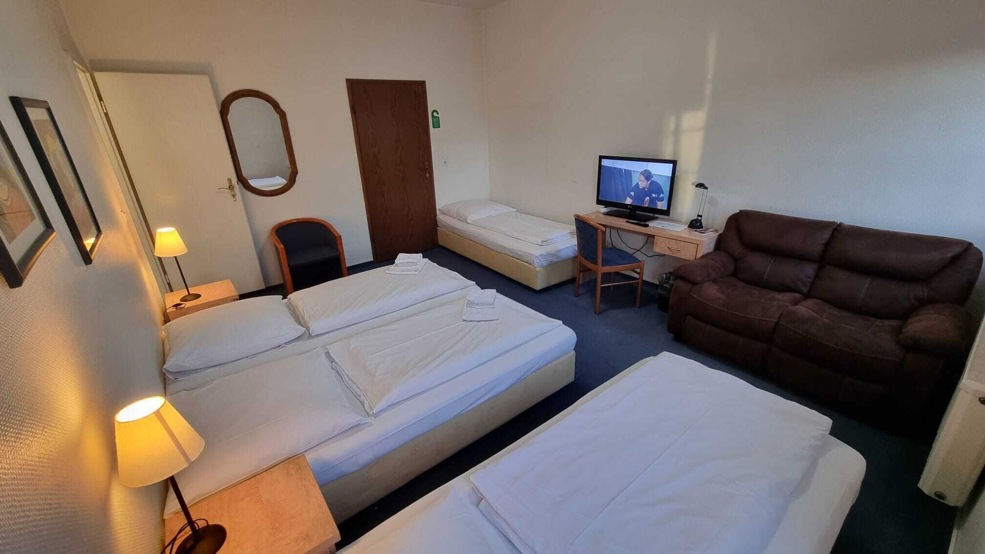 A hotel room with two beds, a desk with a computer, a brown sofa, a chair, a lamp, and a mirror.