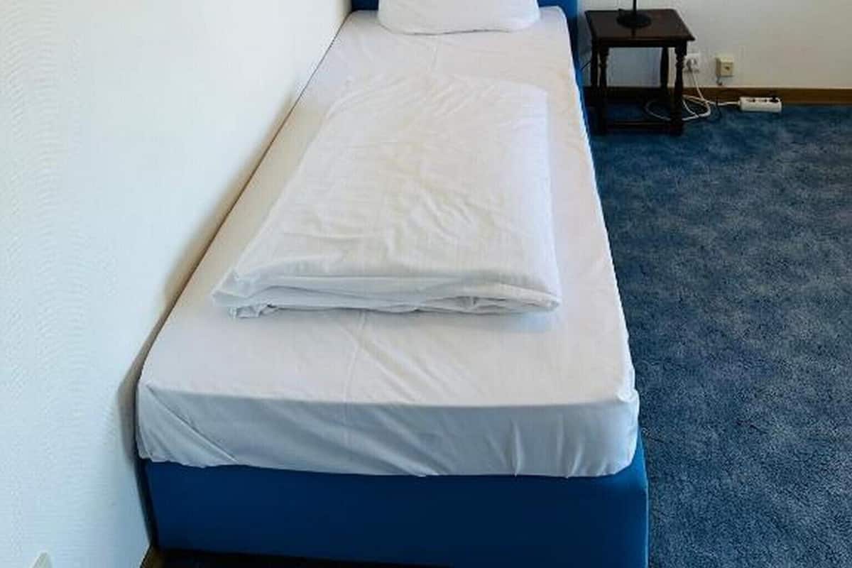 A single bed with a blue headboard and white bedding, a small bedside table with a lamp, and two framed pictures on the wall.