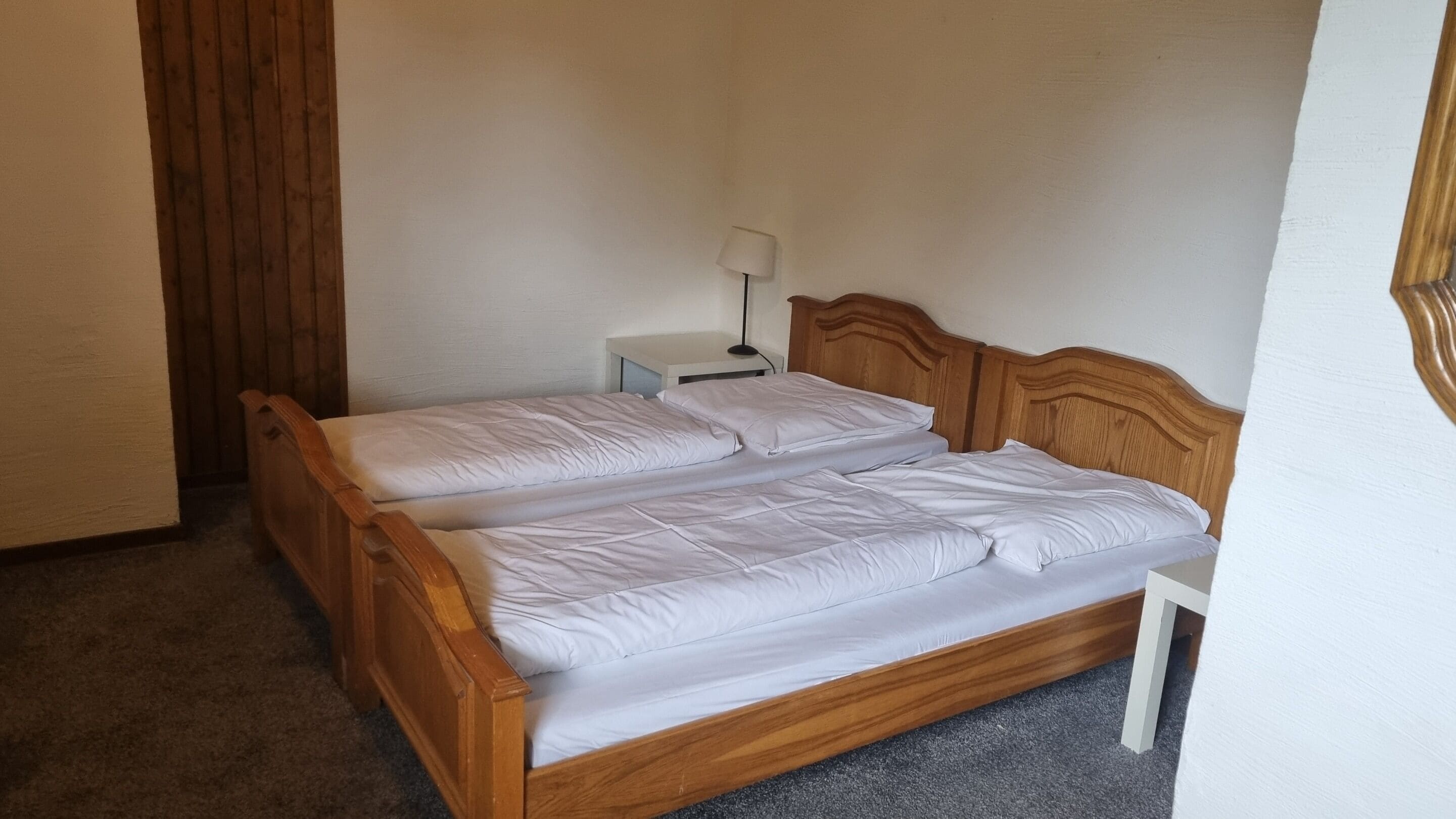 Habitación individual con una cama de una sola plaza, cabecera de madera, una mesita de noche con lámpara y piso cubierto de alfombra.