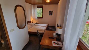 Ein kleiner, kompakter Raum mit zwei Einzelbetten, einem Holztisch mit Lampe, einem kleinen Schreibtisch mit Fernbedienung und einem Fenster mit Blick auf Grünfläche.