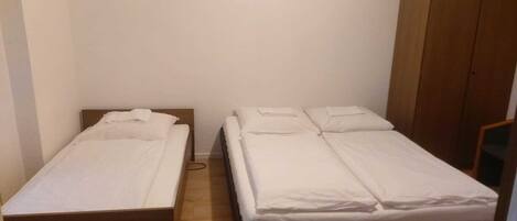 Una habitación pequeña y sencilla con dos camas individuales, un armario de madera y una pared blanca lisa.