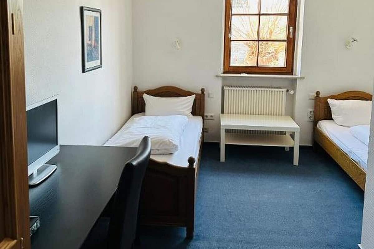 A small, well-lit room with a single bed, a desk, and a chair. There is a window providing natural light and a radiator under the window.