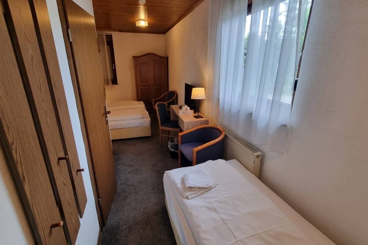 A compact hotel room with a single bed, a chair, a desk, a wardrobe, and a window with curtains.