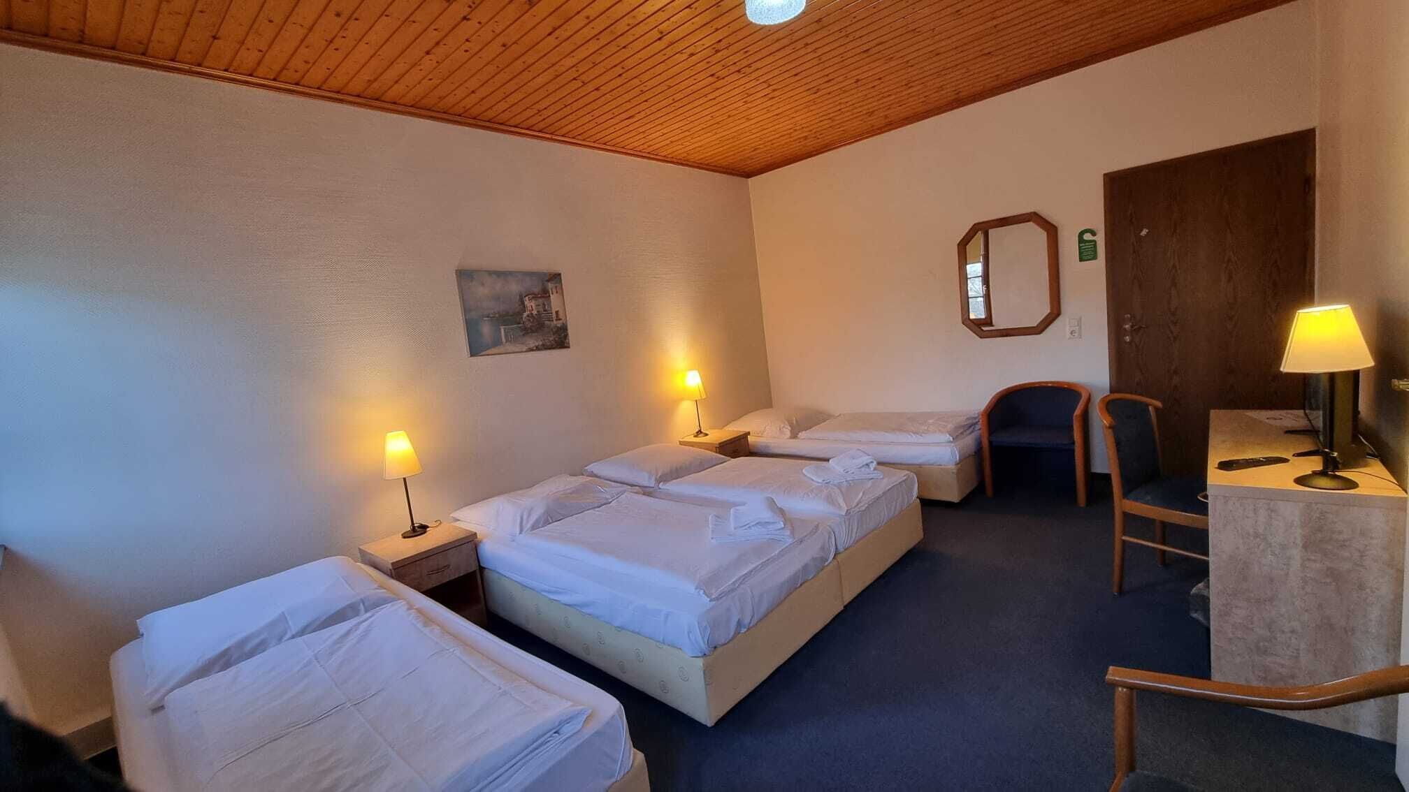 A hotel room with three single beds, a desk with a chair, a wooden ceiling, and a painting on the wall.