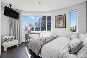 Ein modernes Hotelzimmer mit einem großen Bett, einem Schreibtisch mit Stuhl, einem an der Wand montierten Fernseher und großen Fenstern mit Blick auf die Stadt.