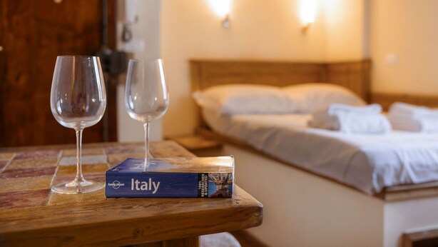 Une table en bois avec deux verres à vin vides et un livre intitulé « Lonely Planet Italie » dans une chambre d’hôtel équipée d’un lit et de lampes fixées au mur.
