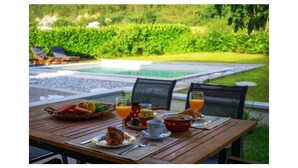 Une table dressée avec des aliments pour le petit-déjeuner, notamment des croissants, des fruits et du jus d’orange, donnant sur une piscine et une végétation luxuriante.