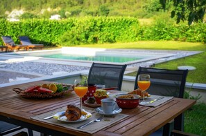 Een gedekte tafel met ontbijtartikelen, waaronder croissants, fruit en sinaasappelsap, met uitzicht op een zwembad en weelderige begroeiing.