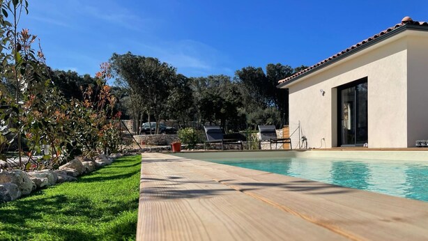 Ein Poolbereich mit Holzterrasse, ein weißes Gebäude mit Ziegeldach und einer grünen Rasenfläche.