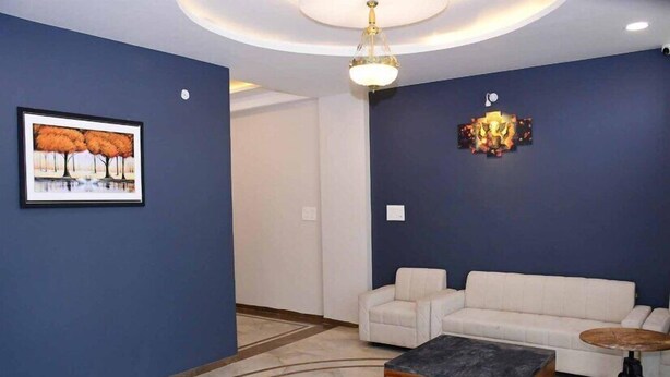 A modern living room with a blue accent wall, a white sofa, a coffee table, and a chandelier. - HOTEL SY21 (Dhar)