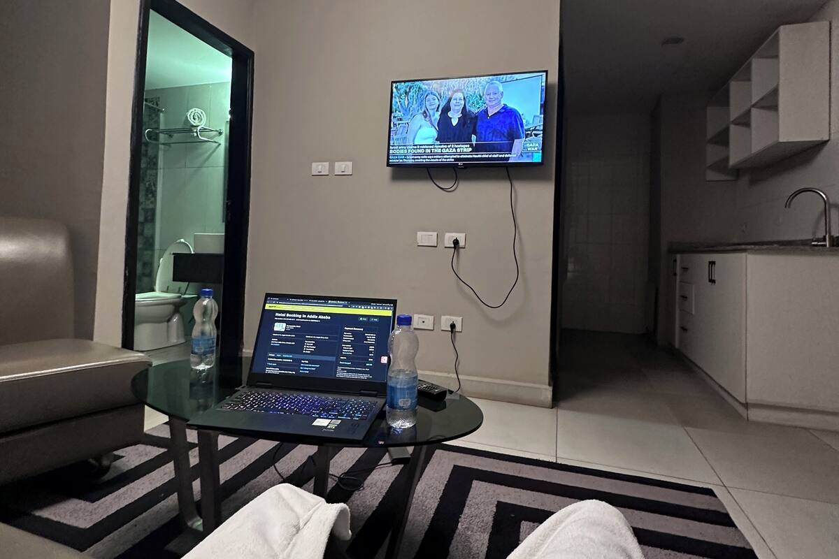 A hotel room with a TV mounted on the wall, a laptop on a glass table, a water bottle, and a bed with a striped rug.