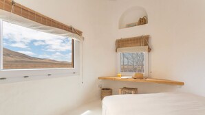 Een slaapkamer met een groot raam, een bed, een houten plank met een hoed en een kleine spiegel.