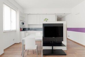 Una cuina moderna amb armaris blancs, un suport de televisió negre i una zona de menjador amb cadires blanques.