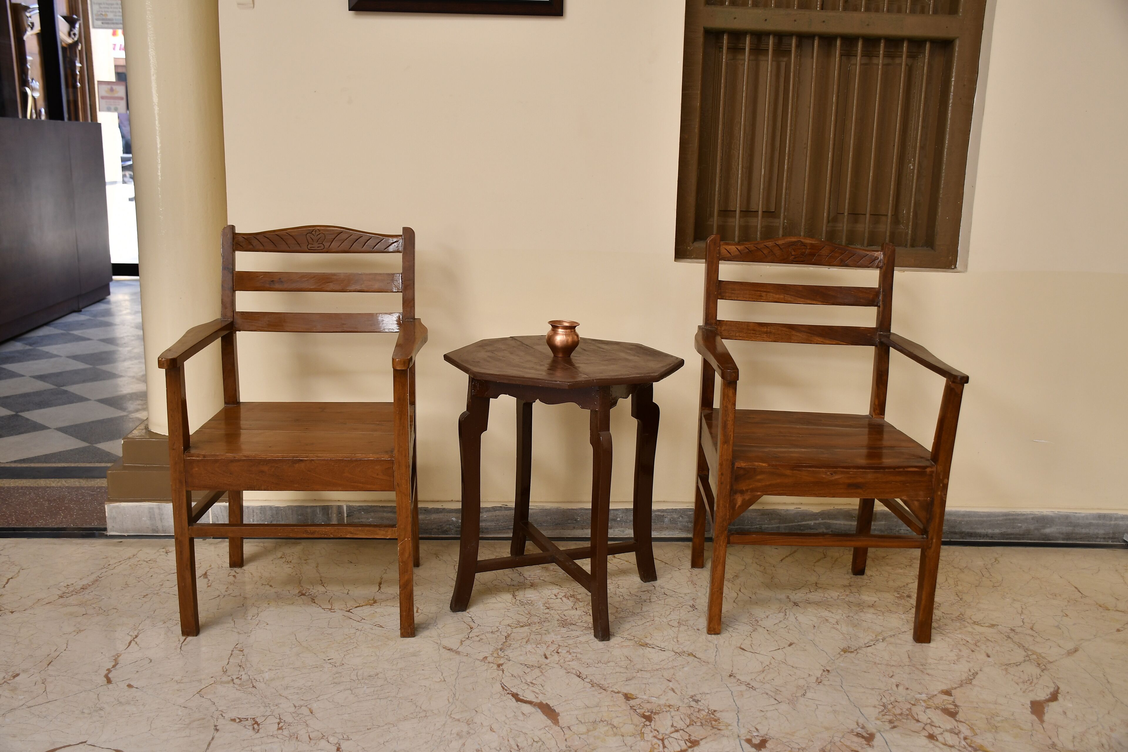 Two wooden chairs and a small table with a copper pot in a hallway with a checkered floor.
