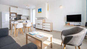 A modern living room with a sofa, a coffee table, a TV, and a dining area with chairs and a table. - Apartment 'Penfret' with Private Terrace, Shared Garden and Wi-Fi (Fouesnant)
