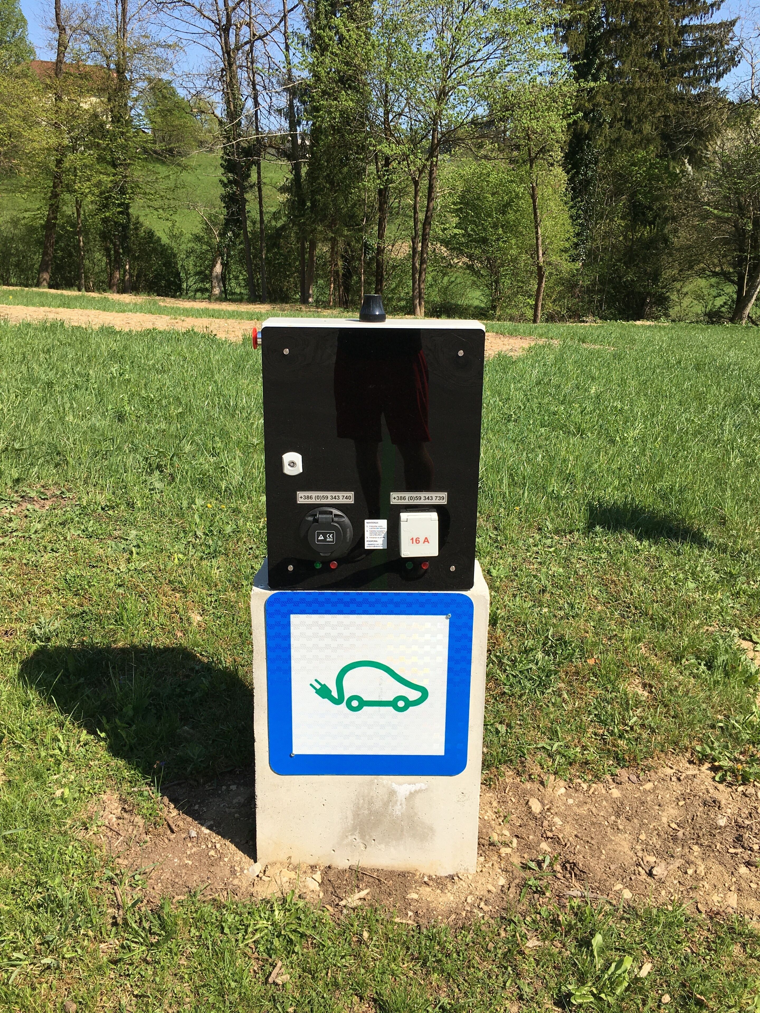 Estación de carga portátil para vehículos eléctricos, con un enchufe y una etiqueta que indica 16 amperios, sobre una base de concreto en un área cubierta de césped.
