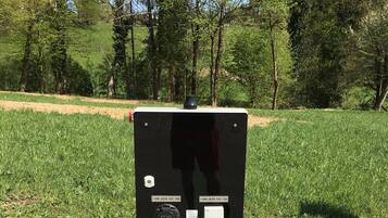 Estación de carga portátil para vehículos eléctricos, con un enchufe y una etiqueta que indica 16 amperios, sobre una base de concreto en un área cubierta de césped.