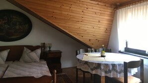A room with a sloped wooden ceiling, a round dining table with a lace tablecloth, a wooden chair, a small potted plant, and a painting on the wall. - Gostišče Atelšek (Rečica ob Savinji)