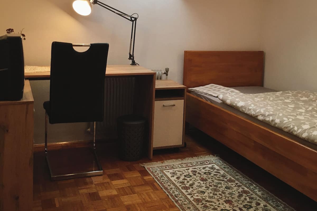 A small, well-lit room with a slanted ceiling, a desk with a chair, a bed, and a radiator.