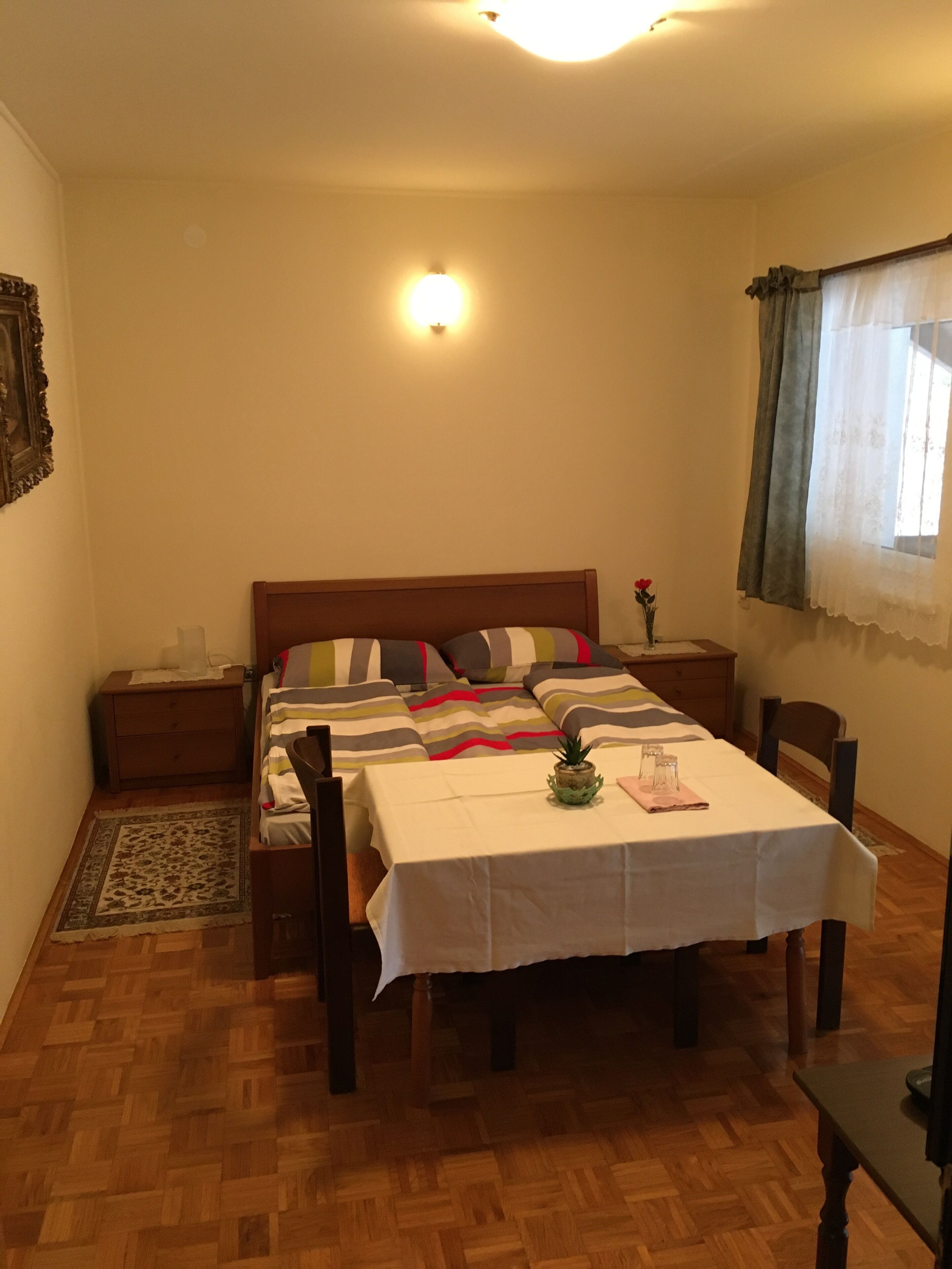 Ein Schlafzimmer mit Bett, Nachttischen, einem Beistelltisch, einem Tisch mit Tischdecke, einem Sessel, Holzboden, Teppich, einem Fenster mit Vorhängen und einer Wandleuchte.