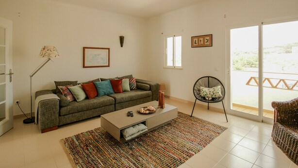 A modern living room with a sofa, coffee table, and a chair. There is a large window and a door leading to an outdoor area.