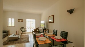 A dining area with a glass table, red placemats, and black chairs. There is a sofa and a chair in the background.
